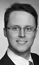 Schwarzweiß-Porträt eines Mannes mit Brille, der ein formelles Outfit trägt. Er lächelt freundlich in die Kamera, während der Hintergrund neutral gehalten ist. Die Bildkomposition vermittelt ein professionelles und selbstbewusstes Auftreten.